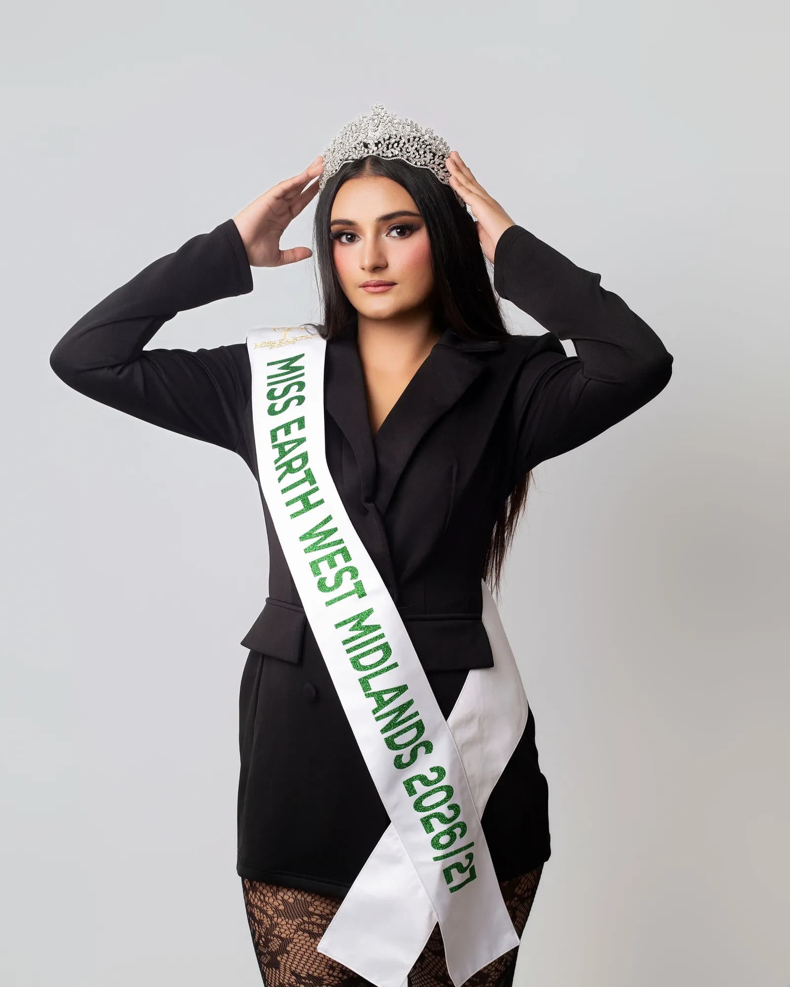 A woman wearing a black jacket, patterned tights, a white sash with green text that reads "MISS EARTH WEST MIDLANDS 2026/27", and a sparkling tiara on her head, with both hands holding the tiara.