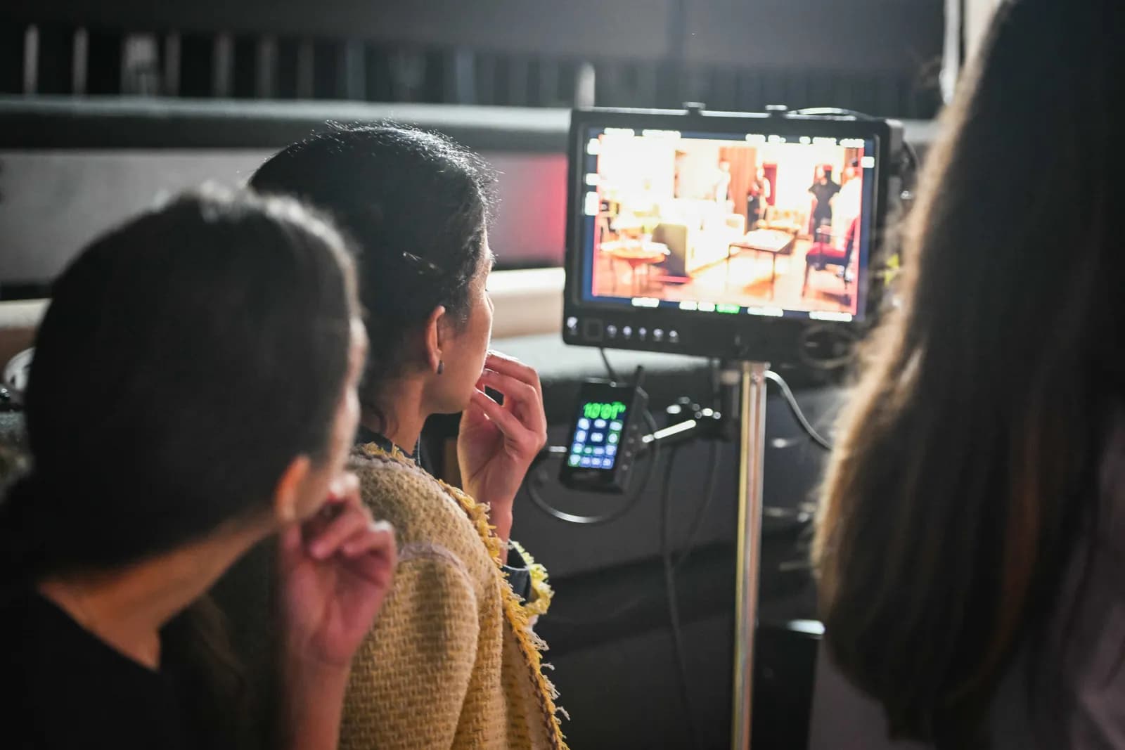 A person with dark hair and a gold tweed jacket looks intently at a production monitor displaying an interior scene with furniture and a standing figure.