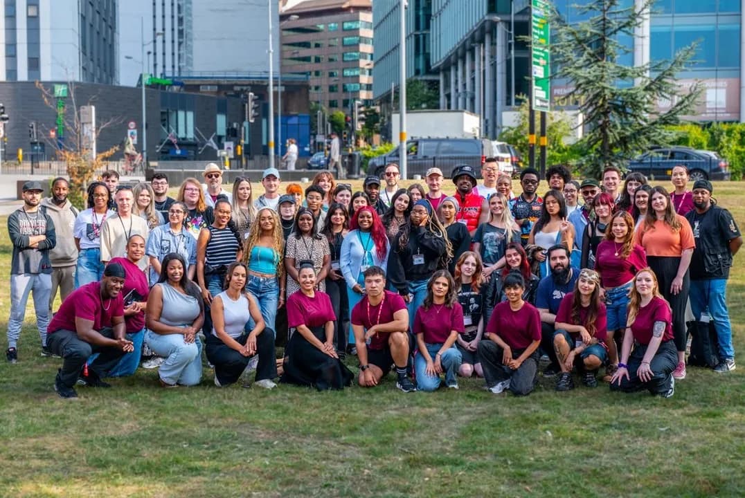 A large group of diverse young adults, some standing and some crouching, pose together on a grassy area in front of city buildings under a bright sky.